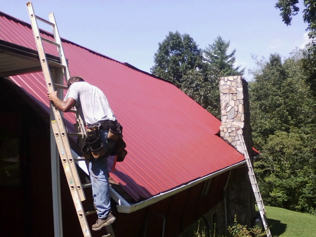 Roofer on a ladder beside a red metal roof and stone chimney — Soffit & Fascia Repair by Roofing Brothers in Nicoma Park